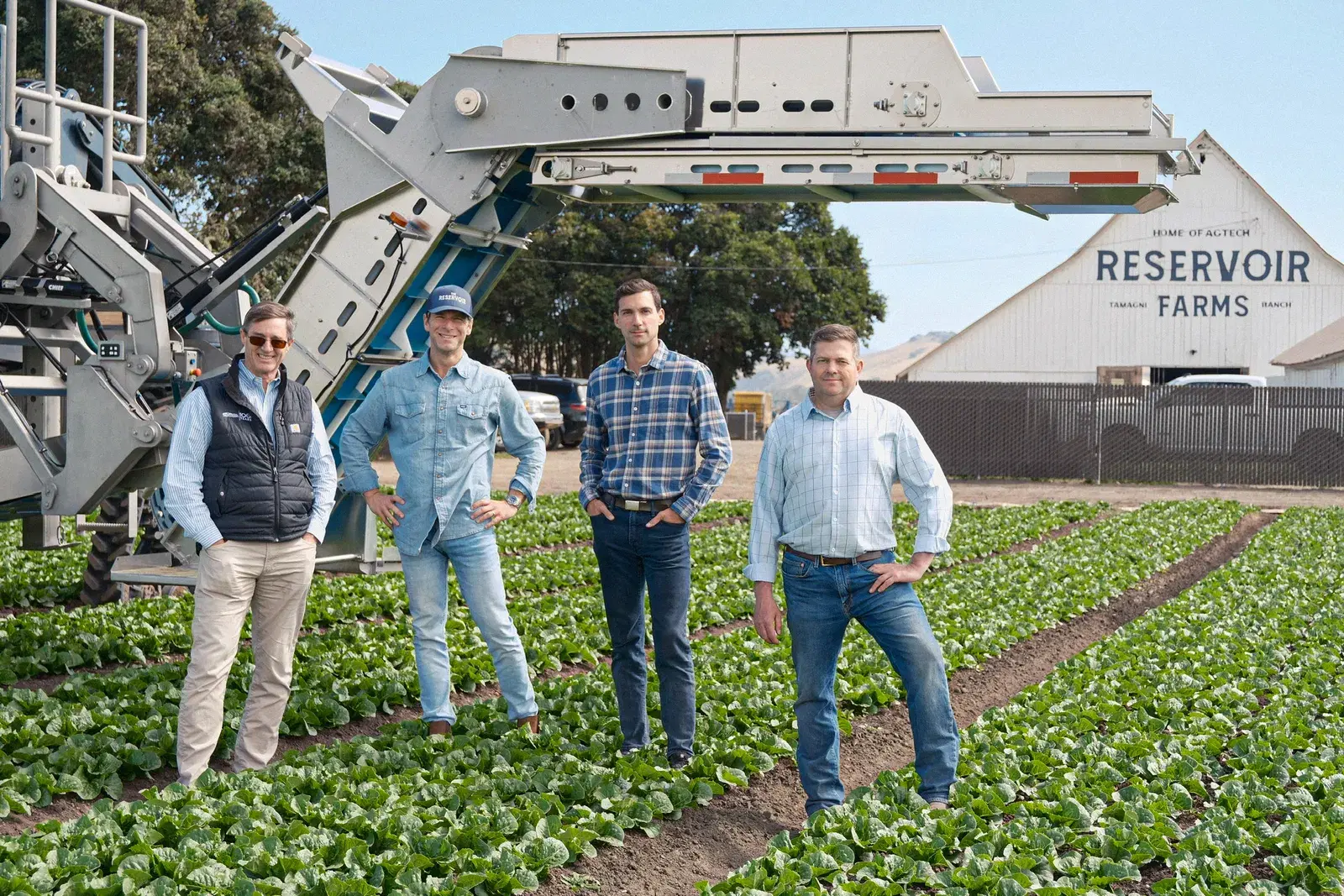 The Sami Robotics and Reservoir Farms teams in a romaine lettuce field at Reservoir Farms in Salinas, with a harvester and the Reservoir Farms barn behind them.